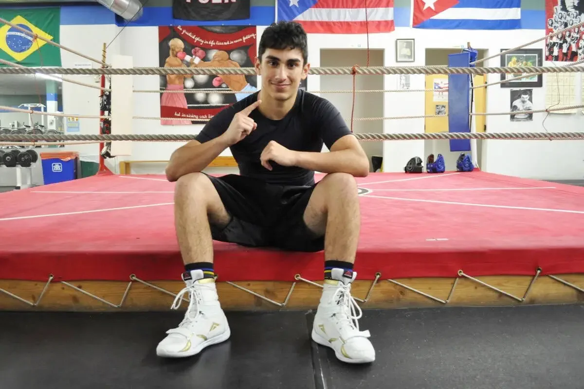 Young man sitting on the edge of a boxing ring in a gym, wearing a black t-shirt, black shorts, white athletic shoes, and colorful ankle socks, with flags, posters, and gym equipment in the background.