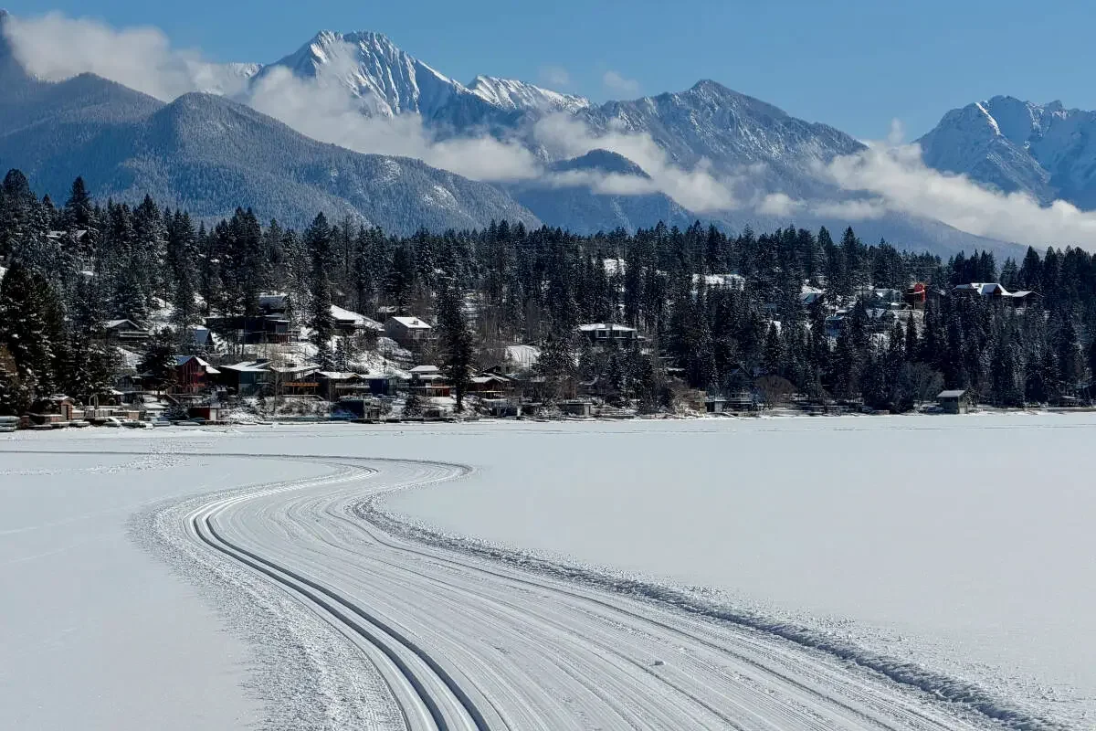 Snow-covered frozen lake with ski tracks, a forested hillside with houses, and snow-capped mountains in the background.