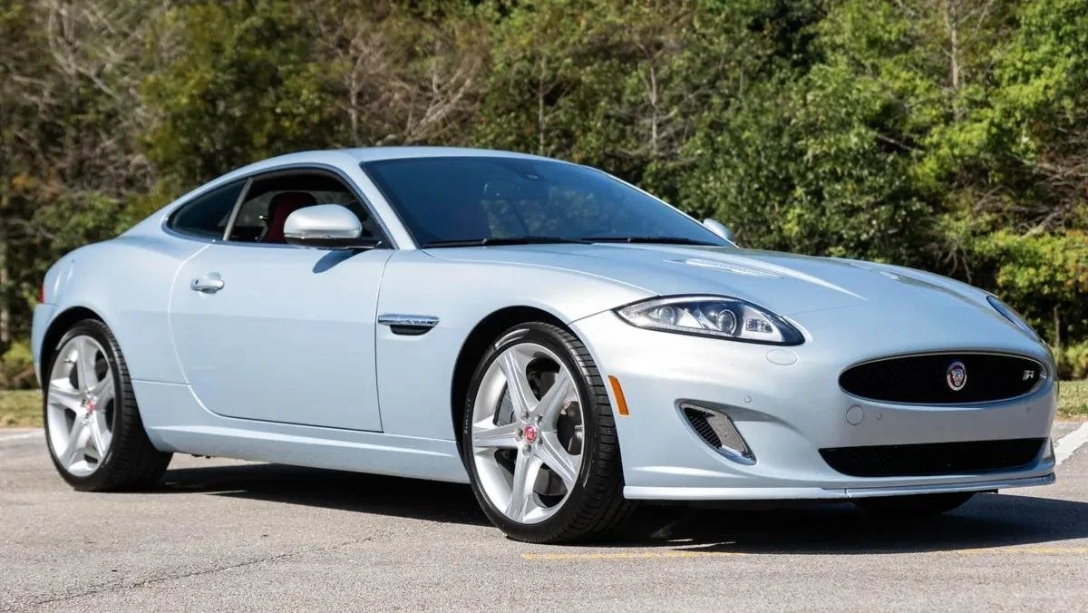 A silver Jaguar coupe car parked on a paved surface with trees in the background.