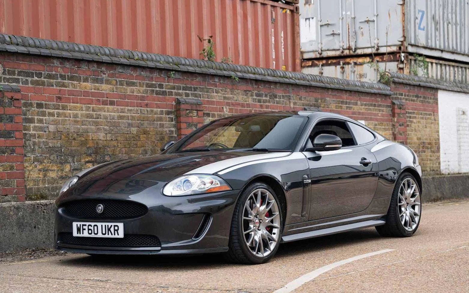 A black Jaguar XK coupe car parked on the street near a brick wall with stacked shipping containers in the background.