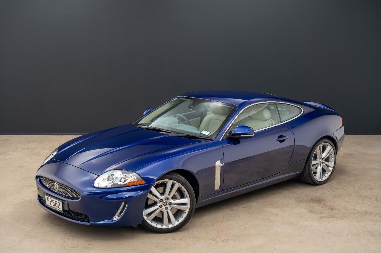 Blue Jaguar XK coupe car parked indoors on a concrete floor with a dark wall background.