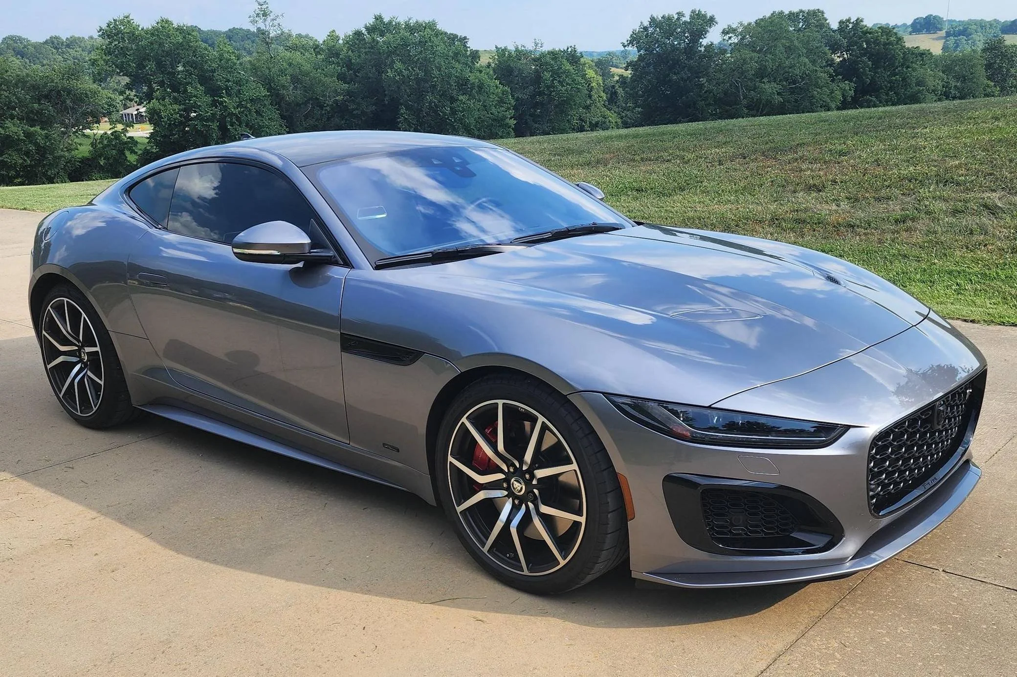A gray sports car parked on a driveway with a grassy hillside and trees in the background.