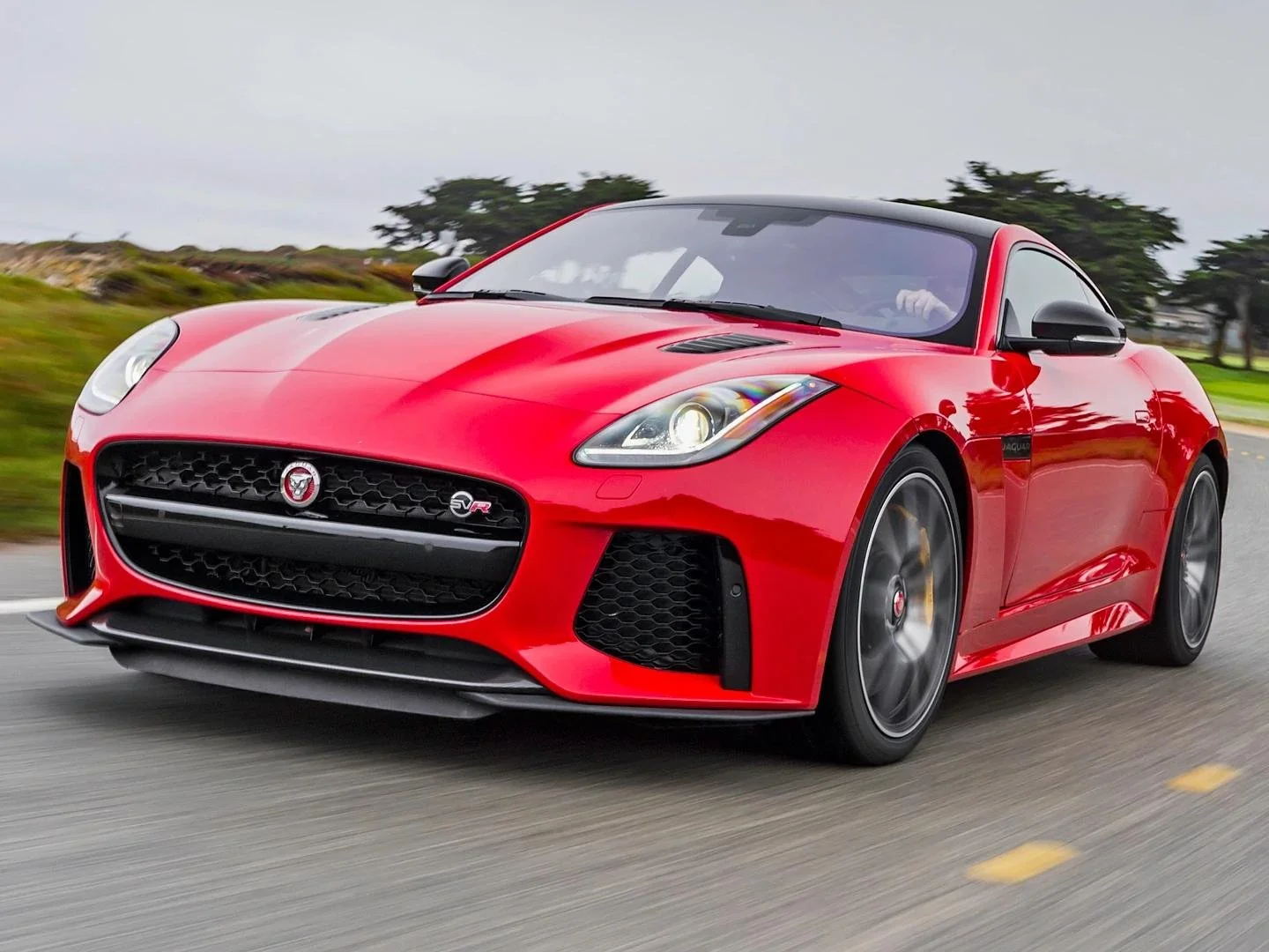 Red Jaguar F-Type sports car driving on a road with a scenic background of trees and sky.