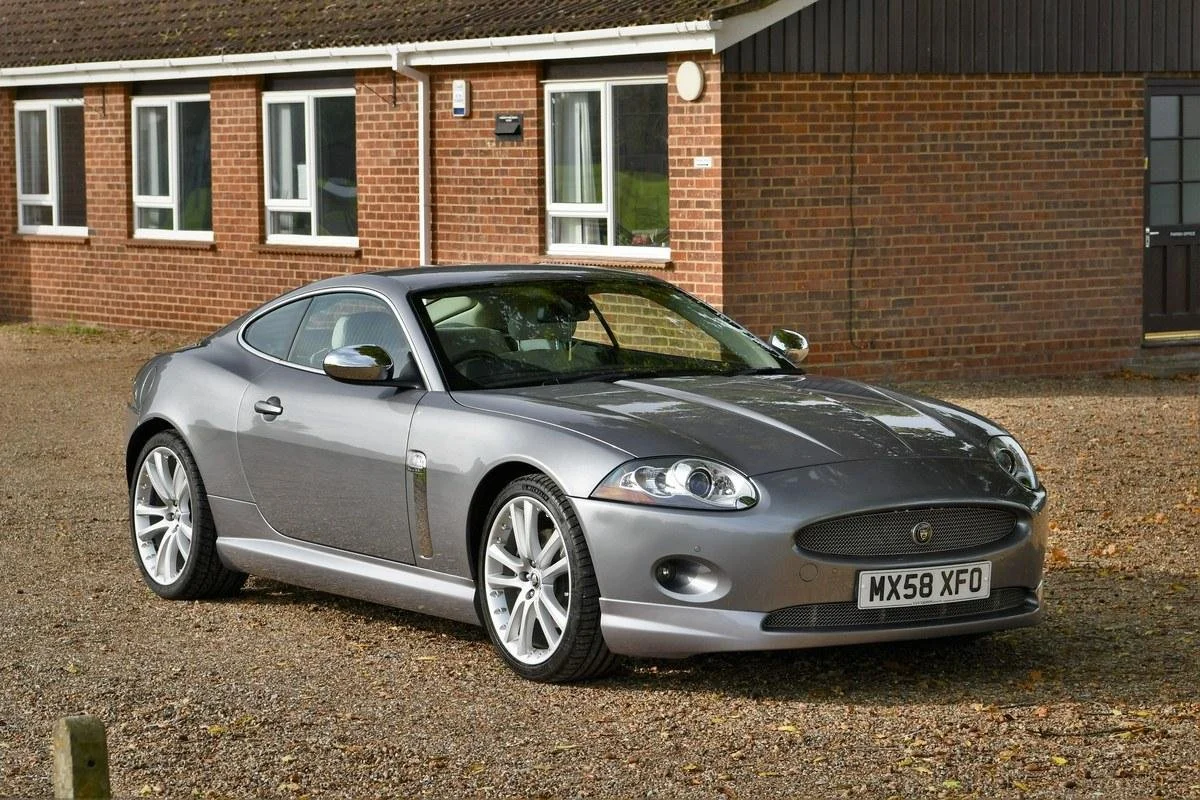 A silver Jaguar XK coupe parked on a gravel driveway in front of a brick building with multiple windows.