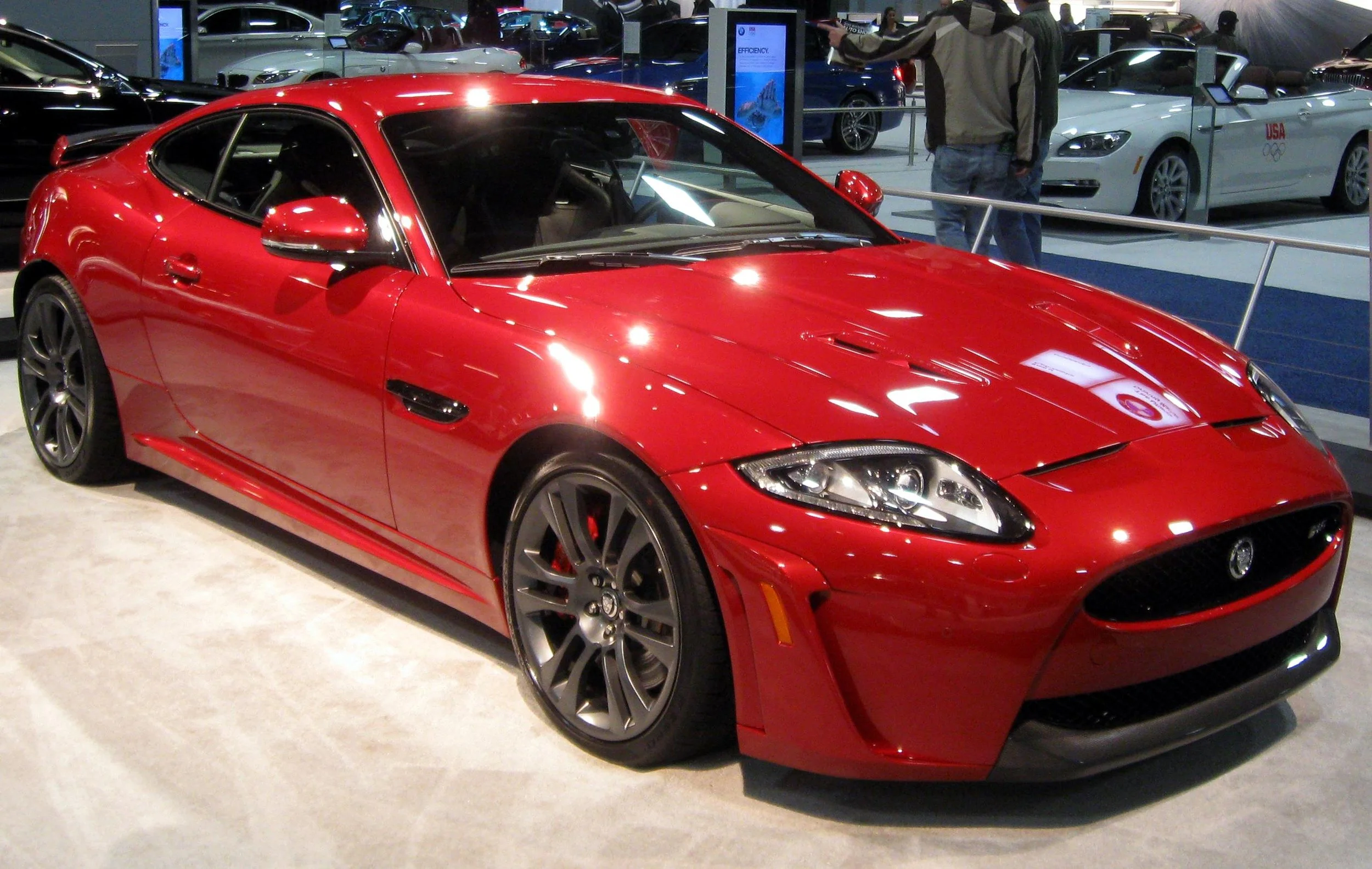 A red sports car on display at an indoor auto show, with other cars and people in the background.