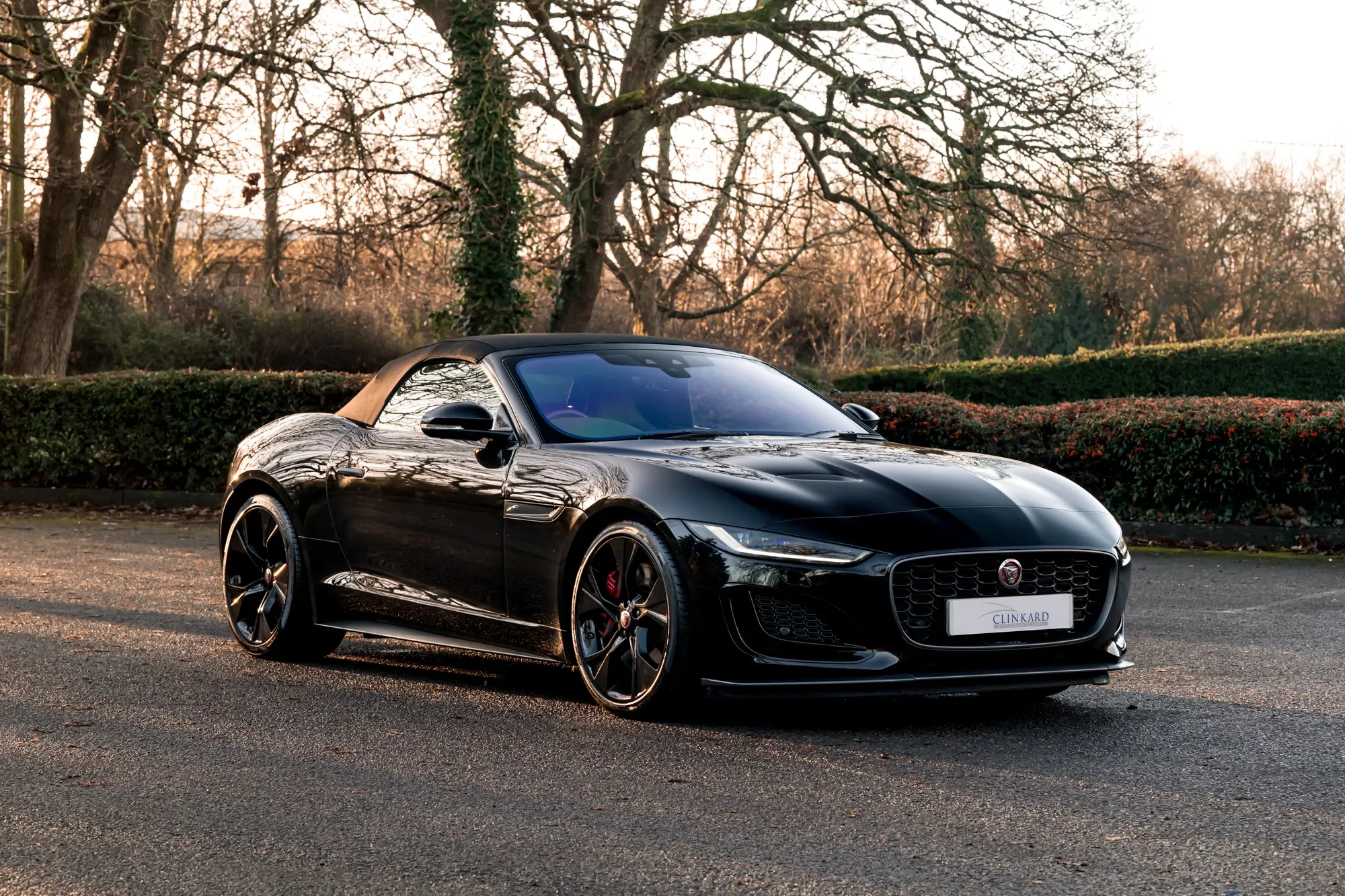 A black Jaguar convertible sports car parked on a paved area with trees and sparse foliage in the background during sunset.