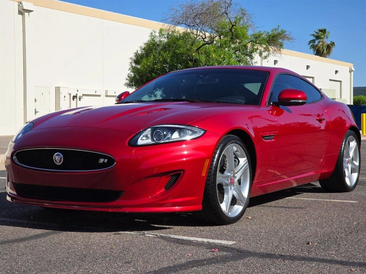 Red Jaguar sports car parked in a parking lot, with trees and a beige building in the background.