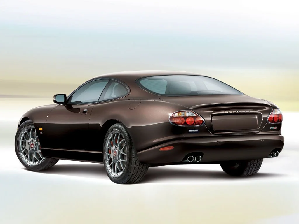 A brown Jaguar XK sports car viewed from the rear left side on a plain background.