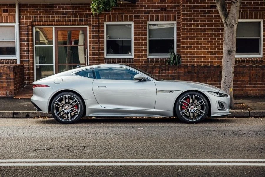 Silver sports car parked beside a brick building and a tree on the sidewalk.