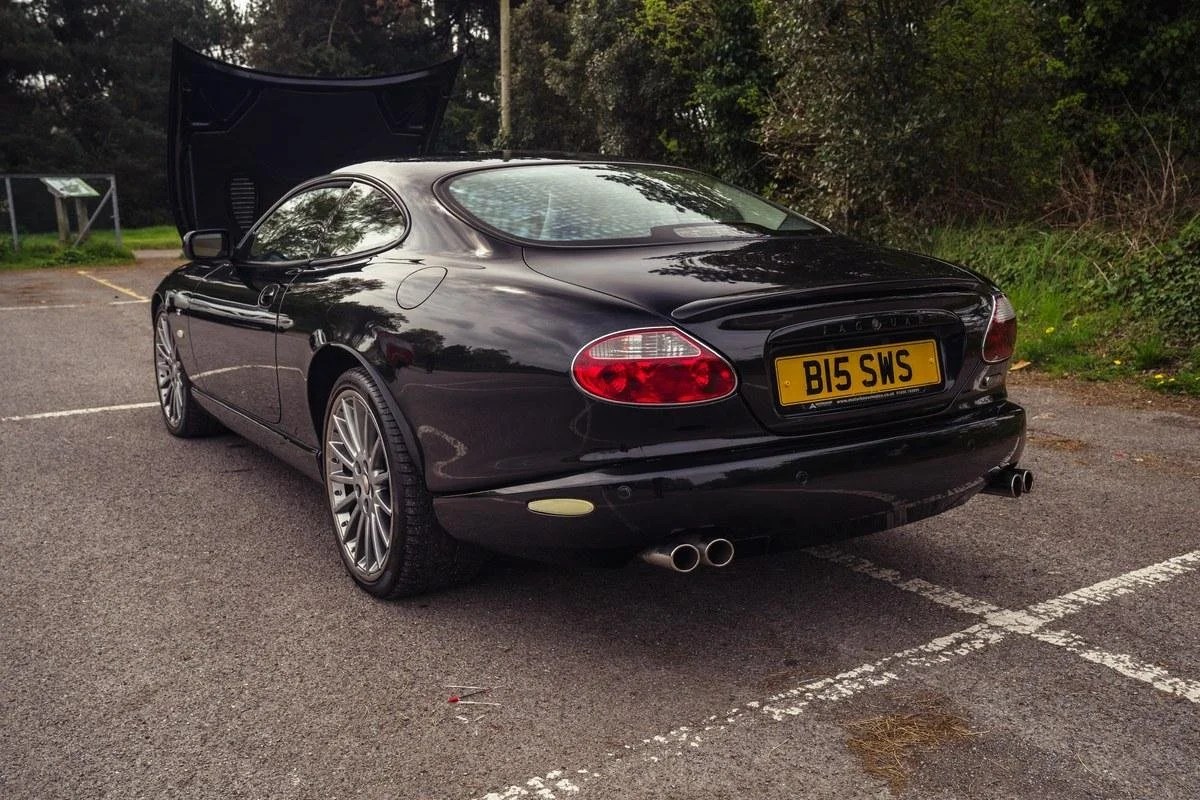 A black Jaguar sports car with dual exhaust pipes parked in a parking lot, with the hood open.