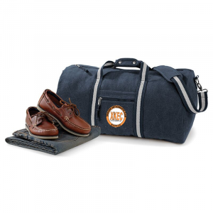 A navy blue duffel bag with a white strap and a brown leather pair of boat shoes placed on a black textured mat.
