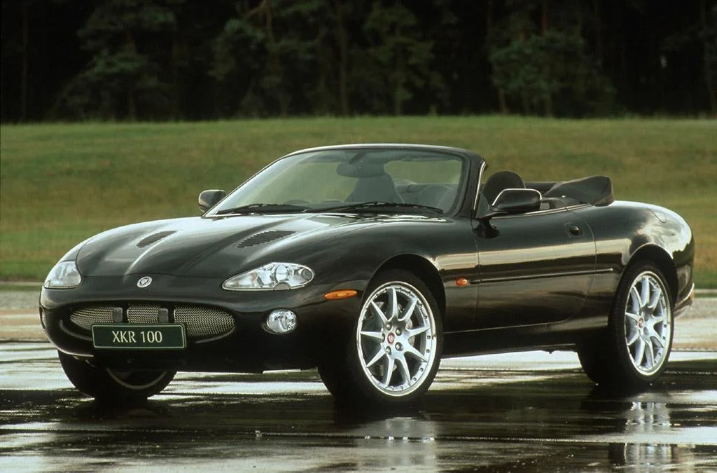 Black convertible sports car with silver wheels parked on a wet surface, greenery in the background.