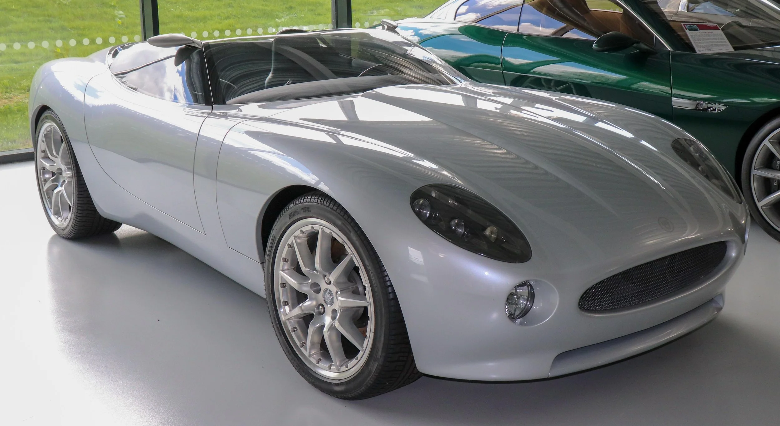 A silver sports car with sleek design and black window tint, displayed indoors on a white surface, with other cars visible in the background.