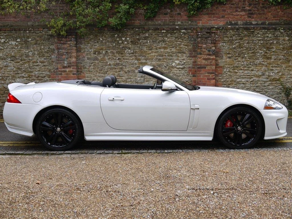A white convertible sports car with a black interior parked on a street, with a brick and stone wall in the background.