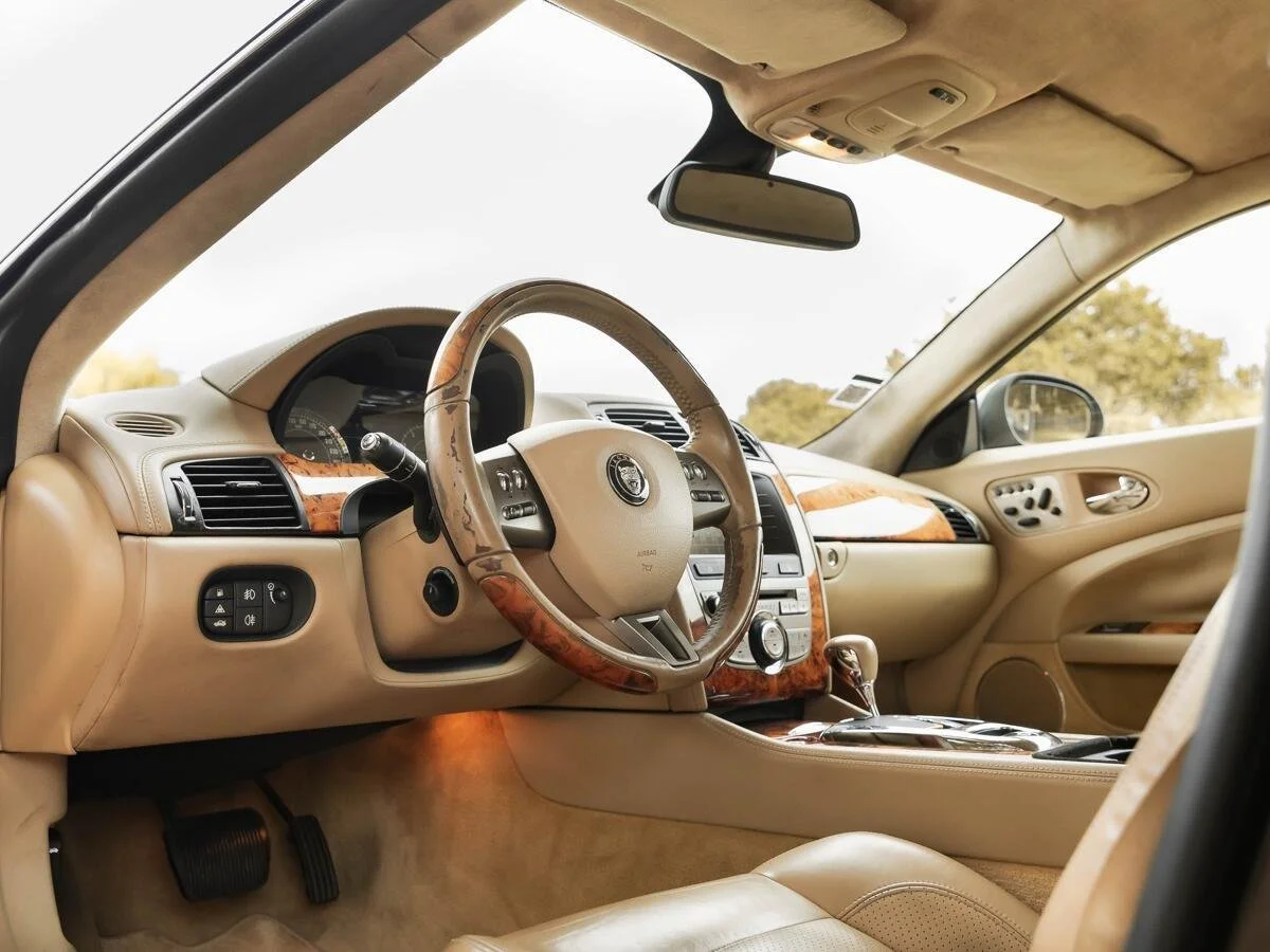 Interior of a luxury car with beige leather seats, wooden trim, and a steering wheel with a logo, dashboard, and gear shift lever.