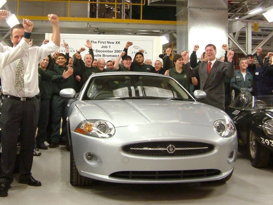 Group of people celebrating around a silver Jaguar XK car in an industrial setting, raising fists in victory.