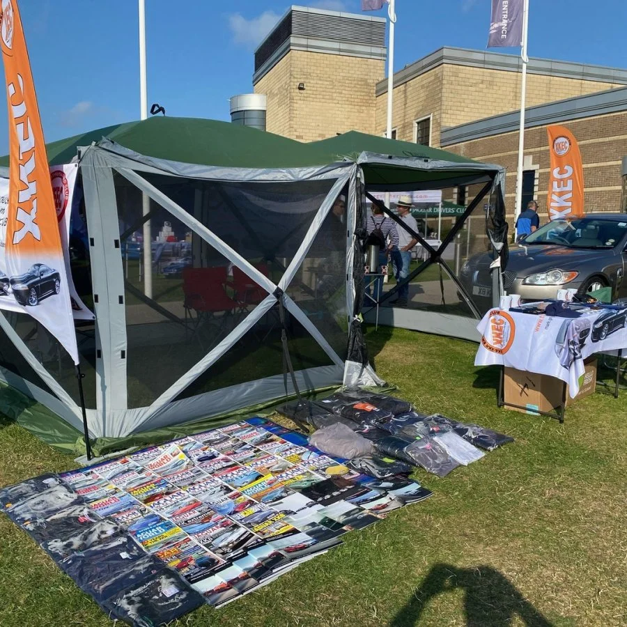 Outdoor car enthusiasts booth at a fair with a tent, car magazines, and branded flags and banners for KWIK E decal and logo, with cars and people in the background.