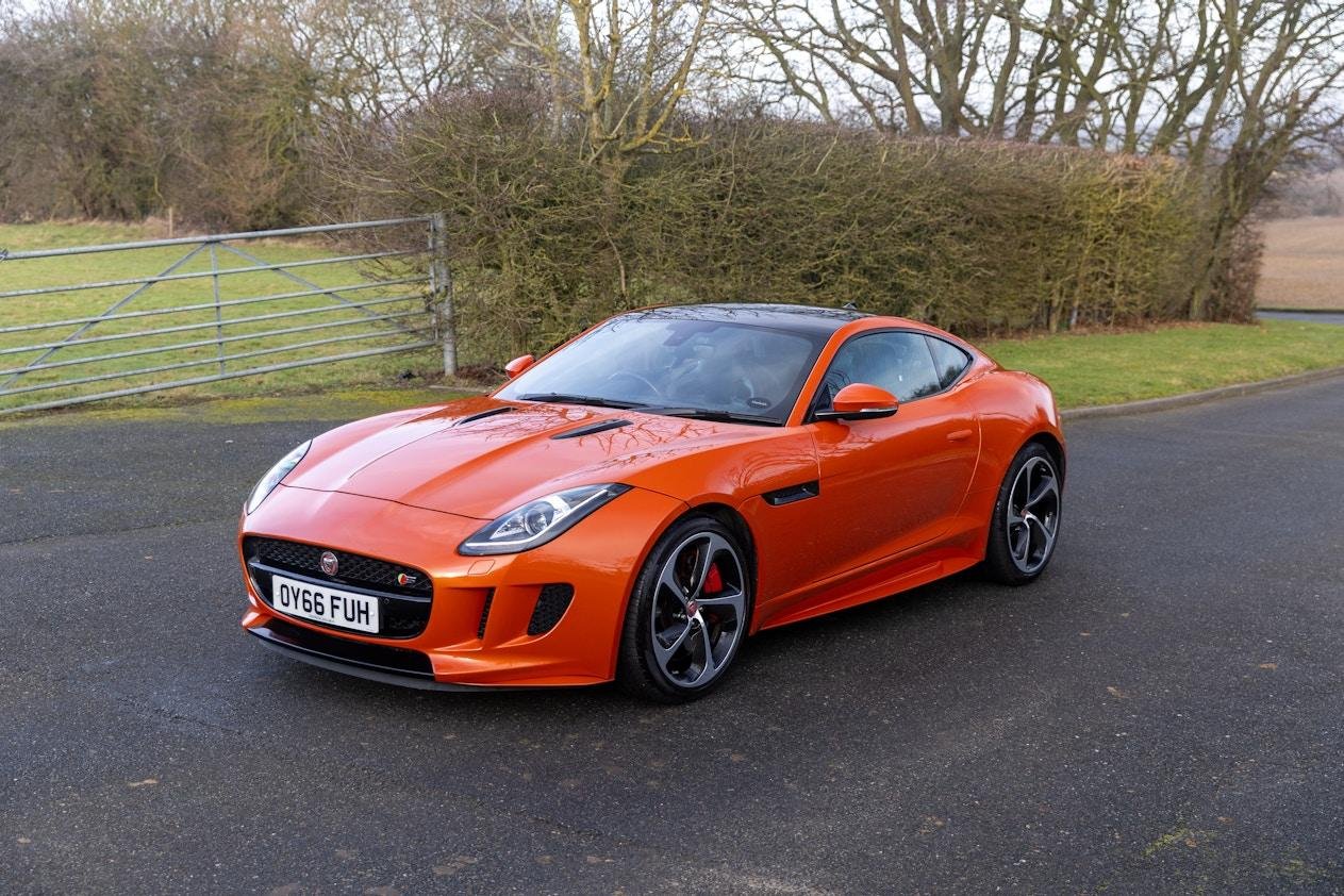 Orange Jaguar sports car parked on a rural road with trees and bushes in the background.