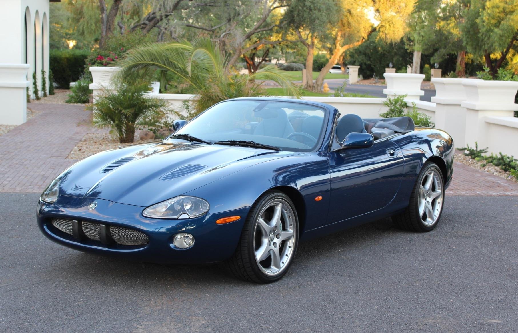 A blue convertible sports car parked on a paved driveway with a landscaped background of trees, shrubs, and a white decorative wall.