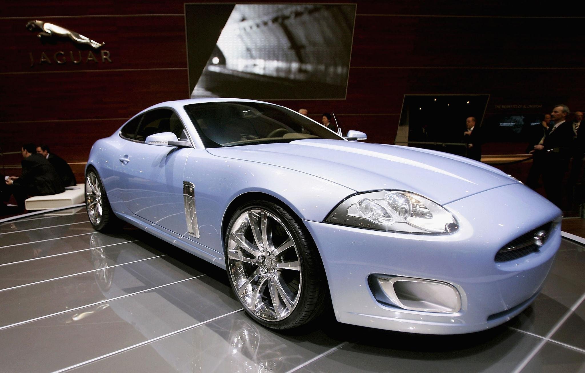 A light blue sports car on display at an auto show, with people in the background and the Jaguar logo on the wall.