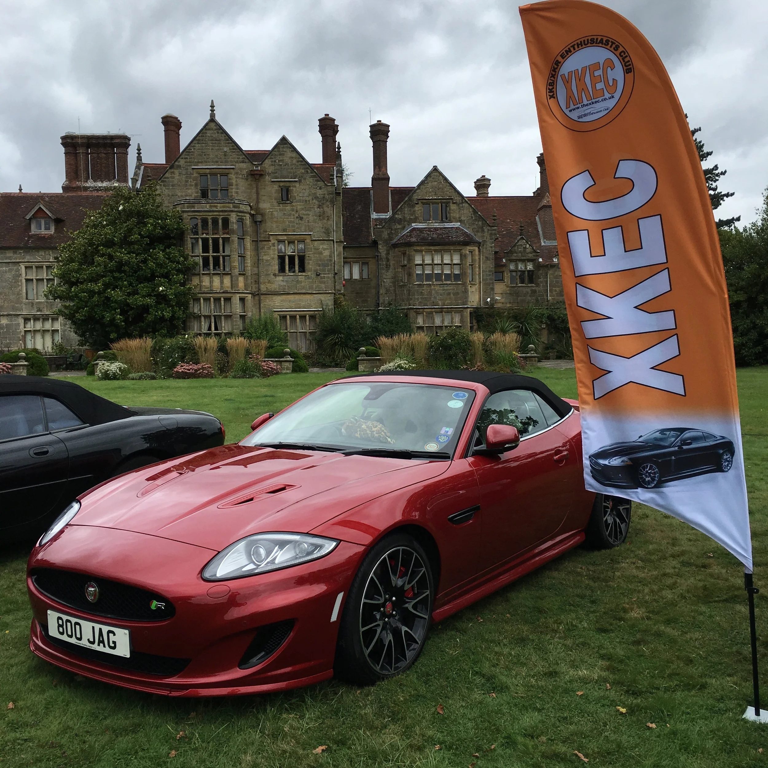 Red Jaguar sports car parked on grass with other cars nearby, and a large orange flag with Vodafone sponsorship on it, in front of an old stone building with multiple chimneys and gabled roofs under an overcast sky.