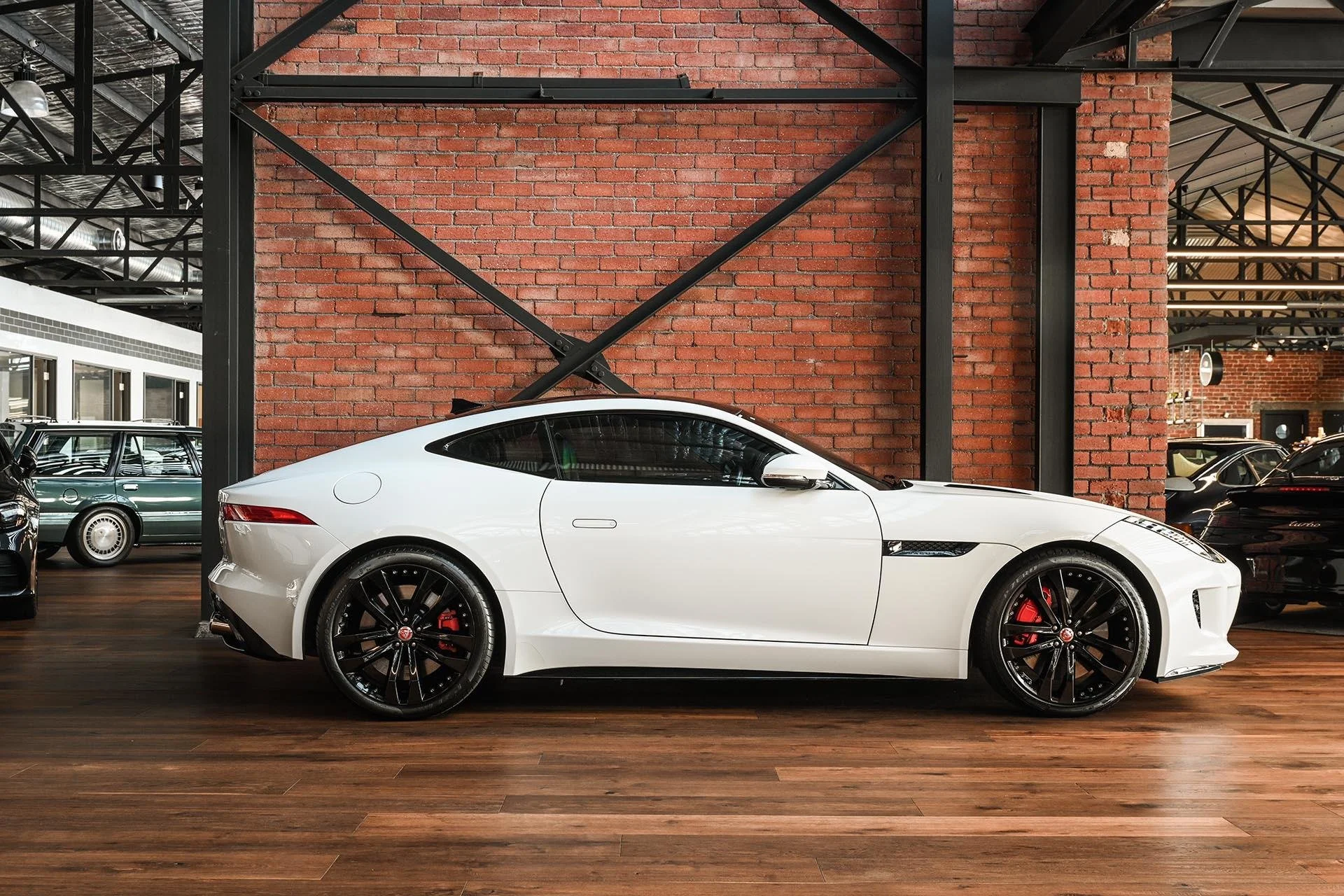 White sports car with black wheels and red brake calipers inside a showroom with a brick wall background and wooden floor.