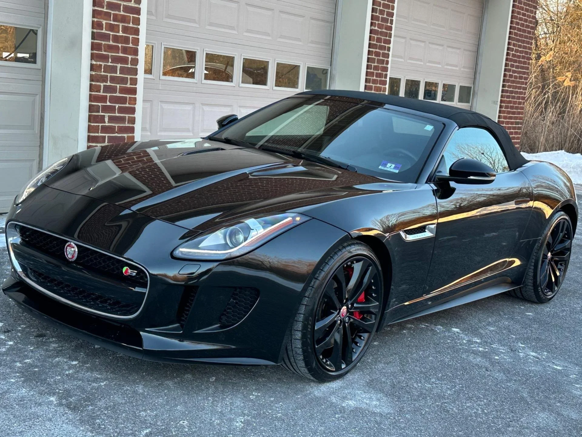 Black Jaguar convertible parked in front of a brick and white garage door, with snow on the ground.