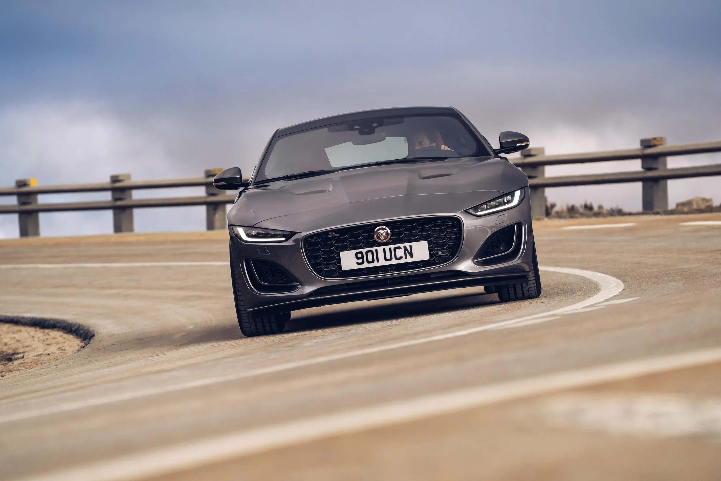 A gray sports car driving on a curved road under a cloudy sky.