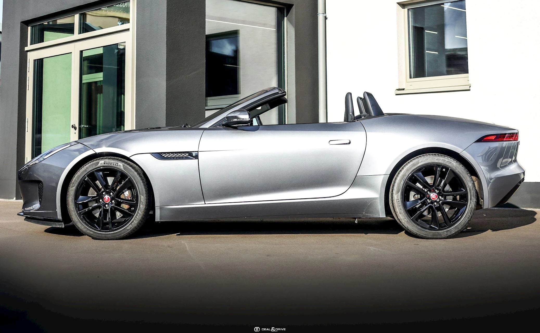 A silver convertible sports car parked in front of a modern building with large windows and a gray and white exterior.
