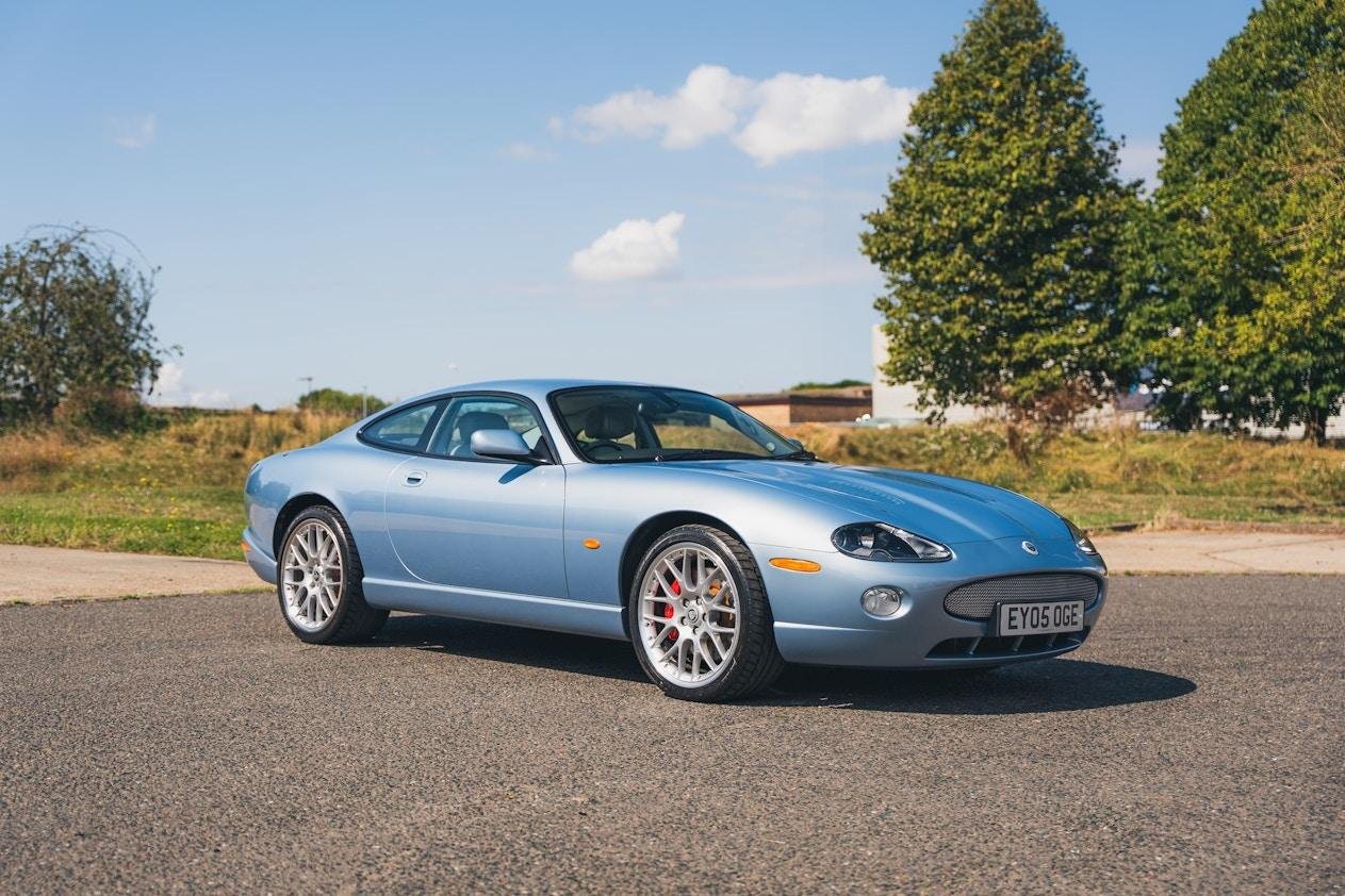 Light blue sports car parked on a paved road with grass and trees in the background under a partly cloudy sky.