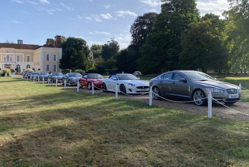 Line of luxury cars parked on grass in front of a large, elegant mansion with trees in the background.