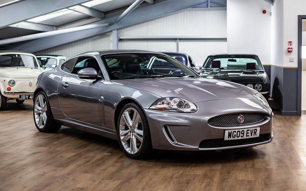 A silver Jaguar sports car parked indoors, surrounded by other vintage vehicles.