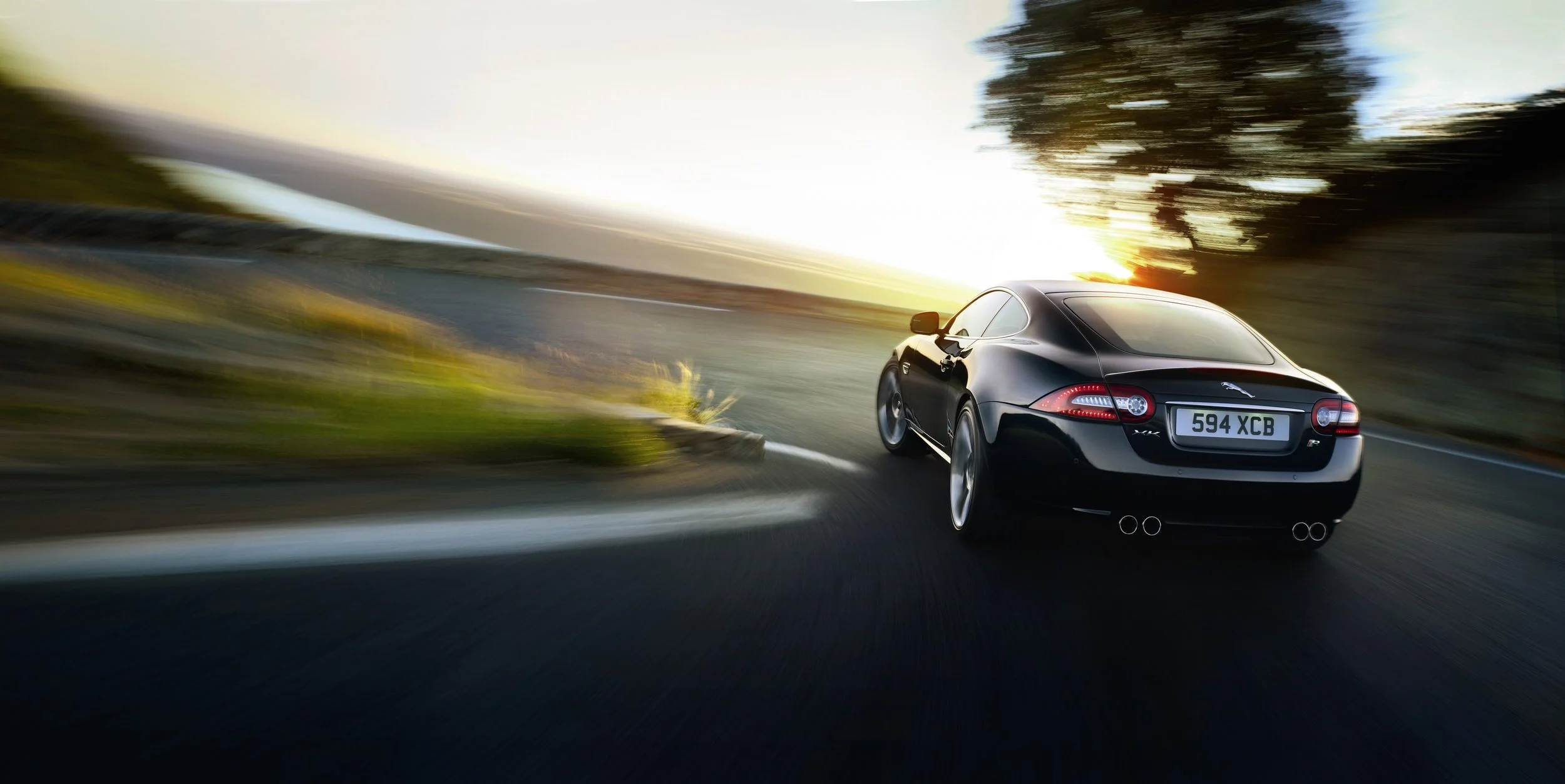 A black sports car driving on a curved road at sunset with a blurred background of trees and sky.