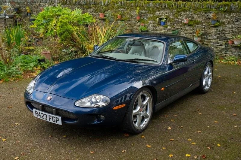 A blue two-door sports car parked on a paved surface in front of a garden with green plants and a brick wall with potted plants. The car has a sleek design and silver wheels.