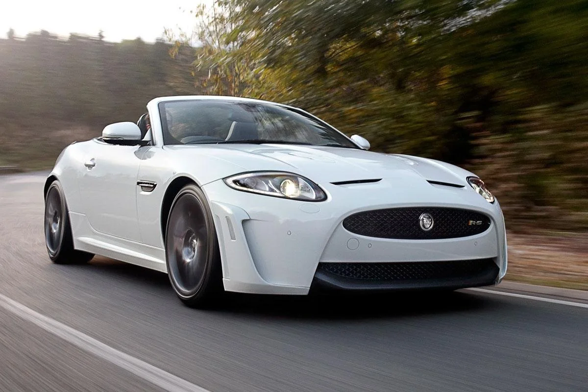 A white Jaguar convertible sports car driving on a road with autumn trees in the background.