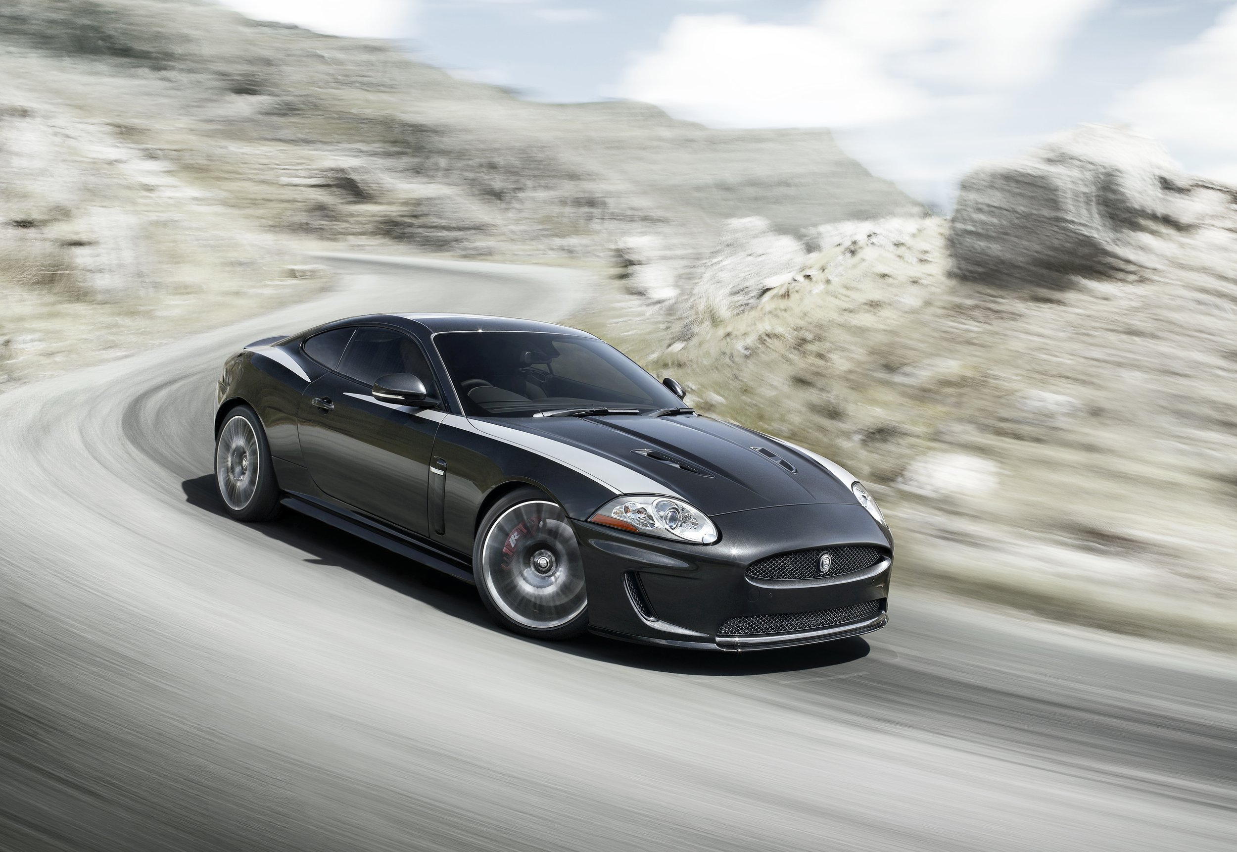A black luxury sports car driving on a winding mountain road with rocky terrain and cloudy sky in the background.