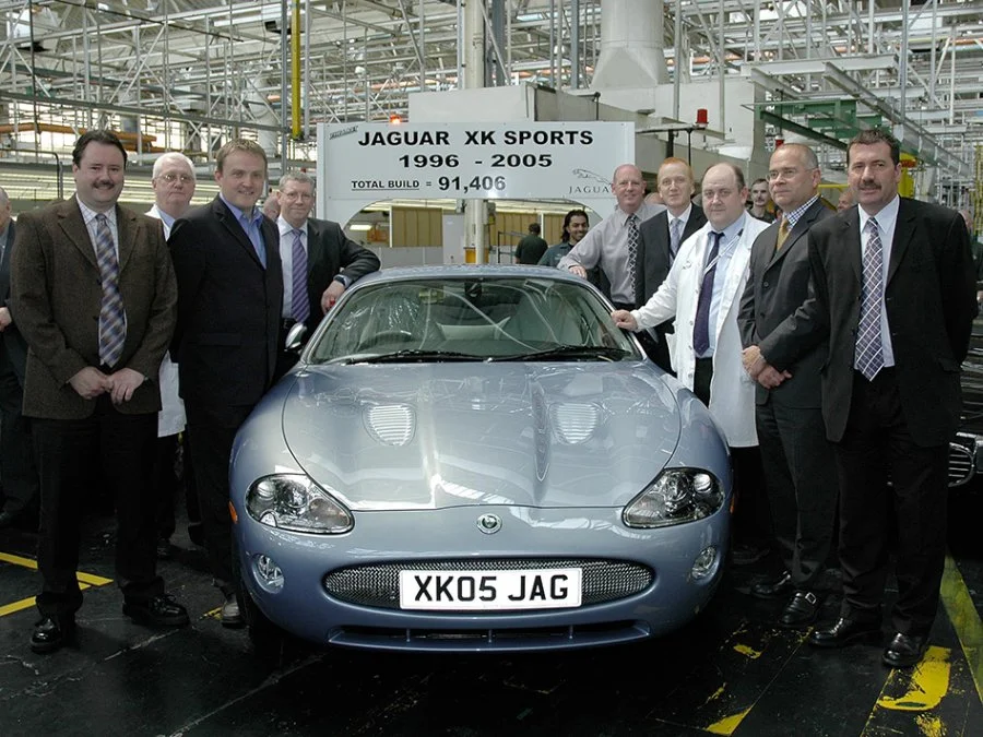 Group of men in formal attire posing around a silver Jaguar XK sports car in an industrial setting. A sign in the background indicates the car was produced from 1996 to 2005, with a total build of 91,406 units. The license plate reads 'XK05 JAG'.