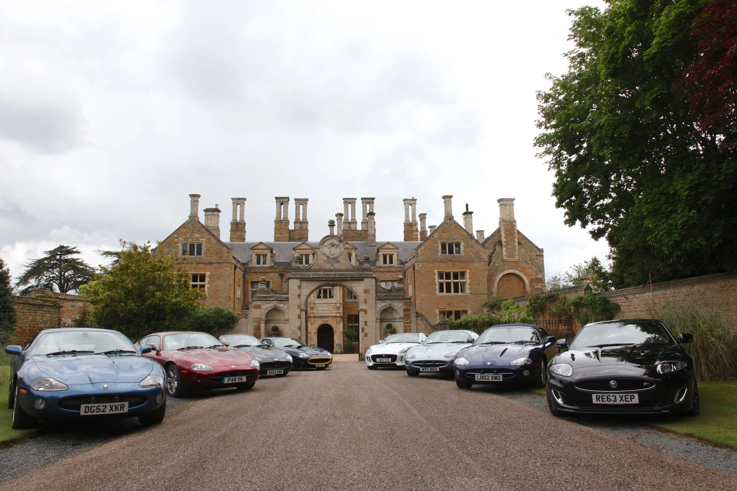 View of a historic stone mansion with multiple chimneys, surrounded by a parking lot filled with sports cars, and framed by green trees under a cloudy sky.