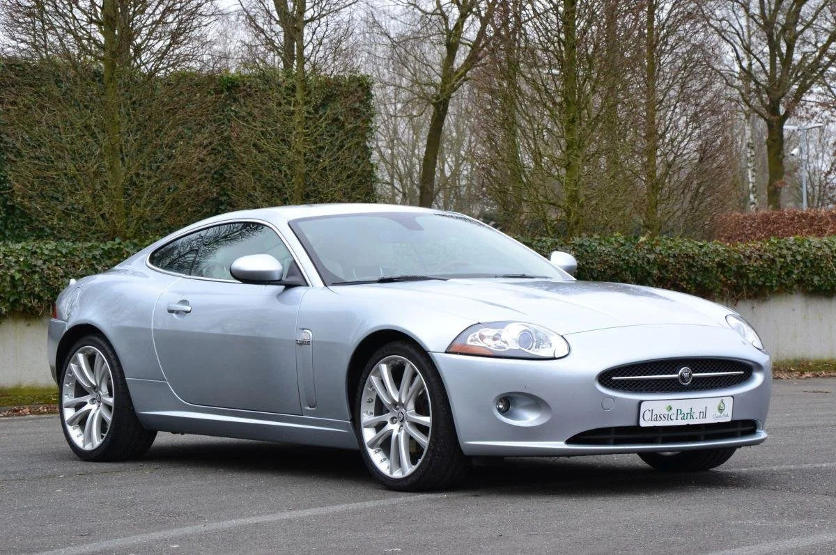 A silver Jaguar XK coupe parked outdoors on a paved surface with trees and bushes in the background.