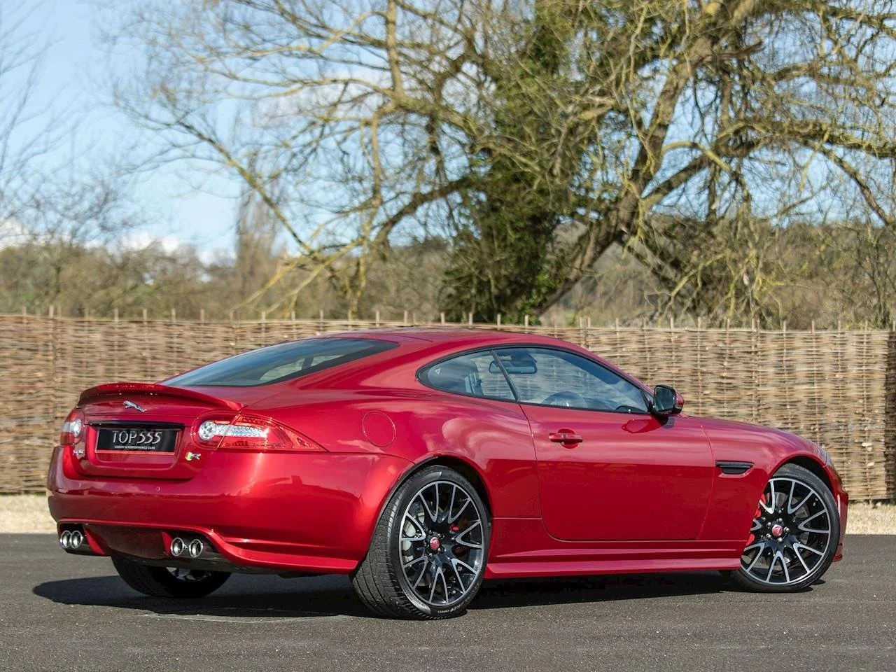 A red sports car parked on a paved surface with a wooden fence and trees in the background under a partly cloudy sky.