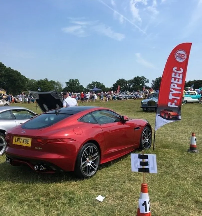 A red sports car on display at a car show on a grassy field, with a crowd of people, vintage cars, and a red flag with a car logo and the word 'E-TYPE' on it.
