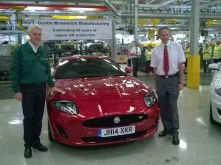 Two men standing on either side of a red Jaguar XK sports car inside a manufacturing plant. The plant has a banner in the background celebrating 66 years of Jaguar XK production at Castle Bromwich.