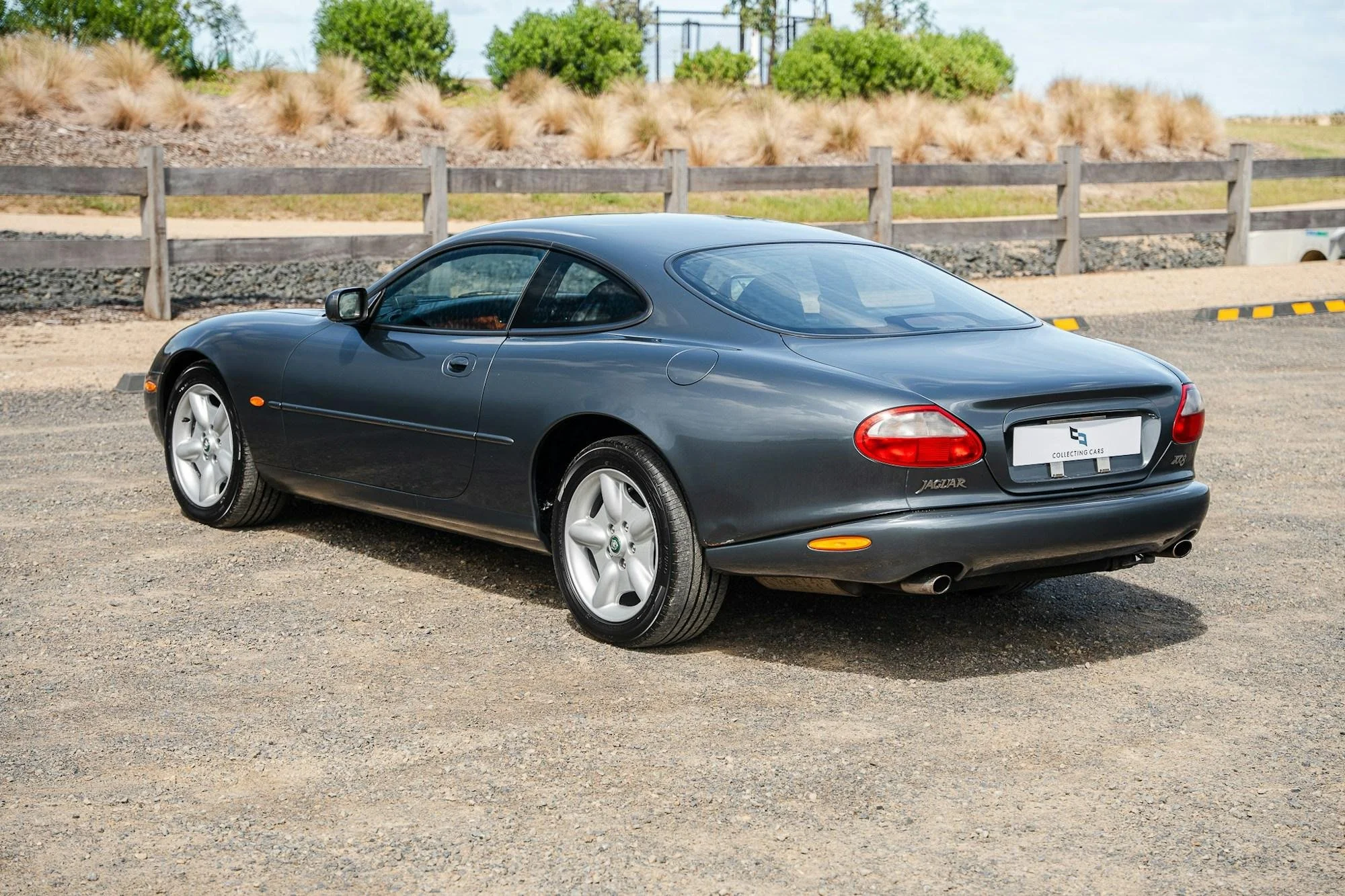A dark gray Jaguar XK sports car parked on a gravel lot with a wooden fence, tall grass, shrubs, and electrical towers in the background under a clear blue sky.