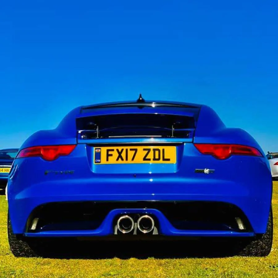 Back of a blue sports car with a yellow UK license plate, dual exhaust pipes, and a rear spoiler, parked on grass under a clear blue sky.