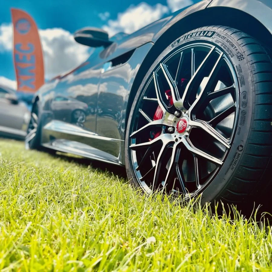 Close-up of a black sports car with a large wheel and Michelin tire, parked on a grassy field under a partly cloudy sky.