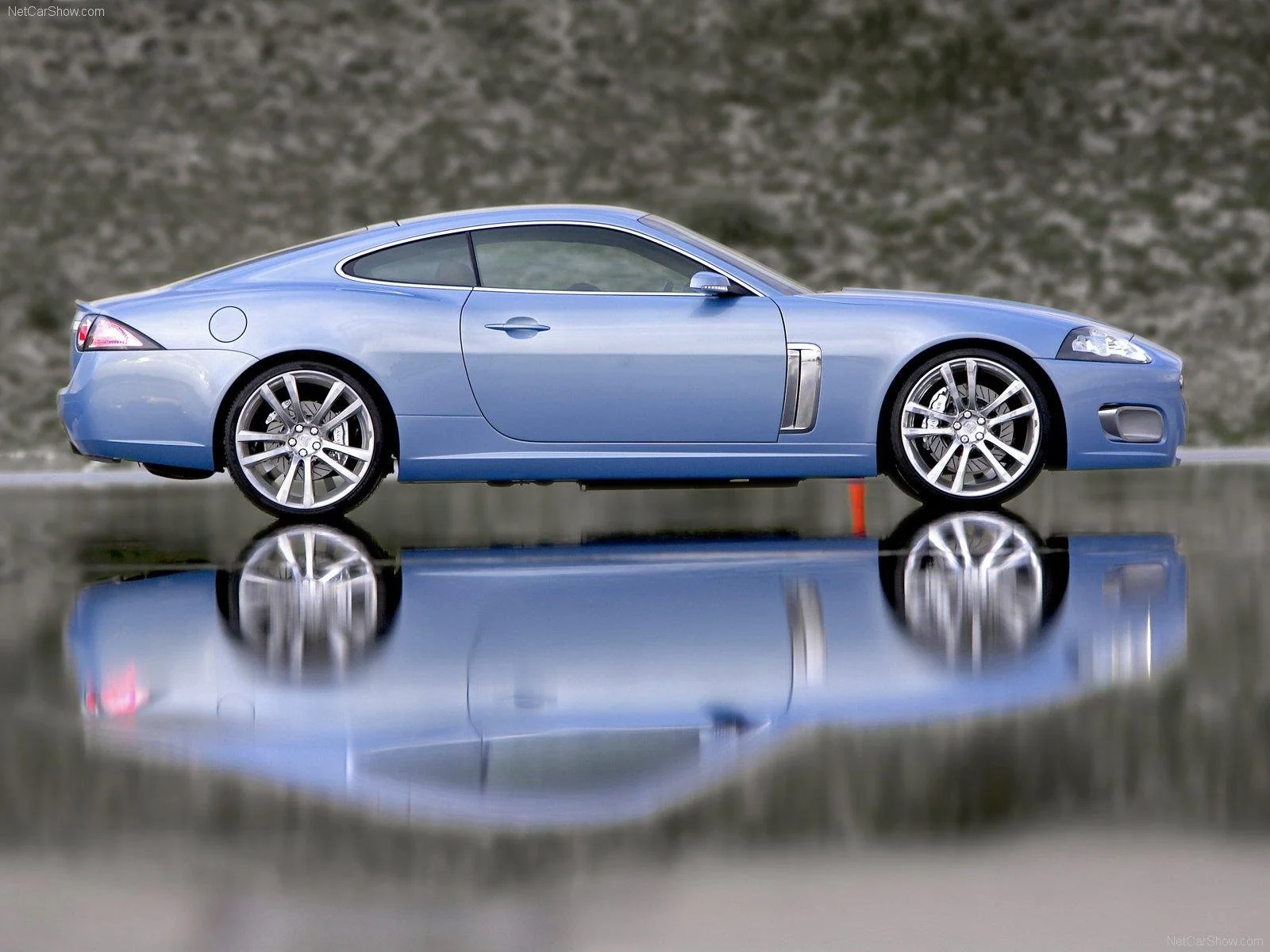 A light blue sports car, parked on a reflective surface with a blurred background. The car has silver rims, a sleek design, and tinted windows.