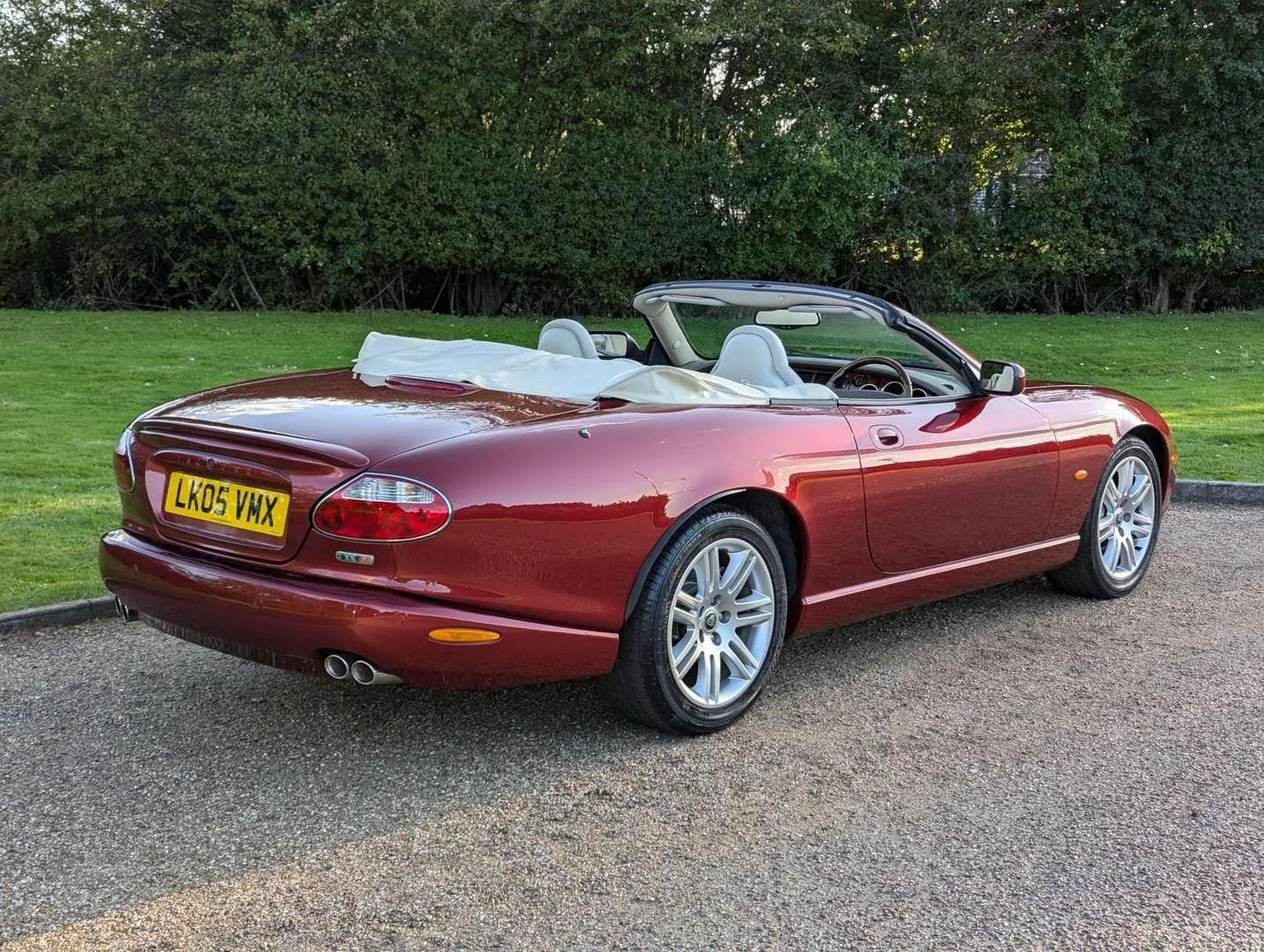 Red convertible car parked on a gravel driveway with a grassy area and trees in the background, soft sunlight.