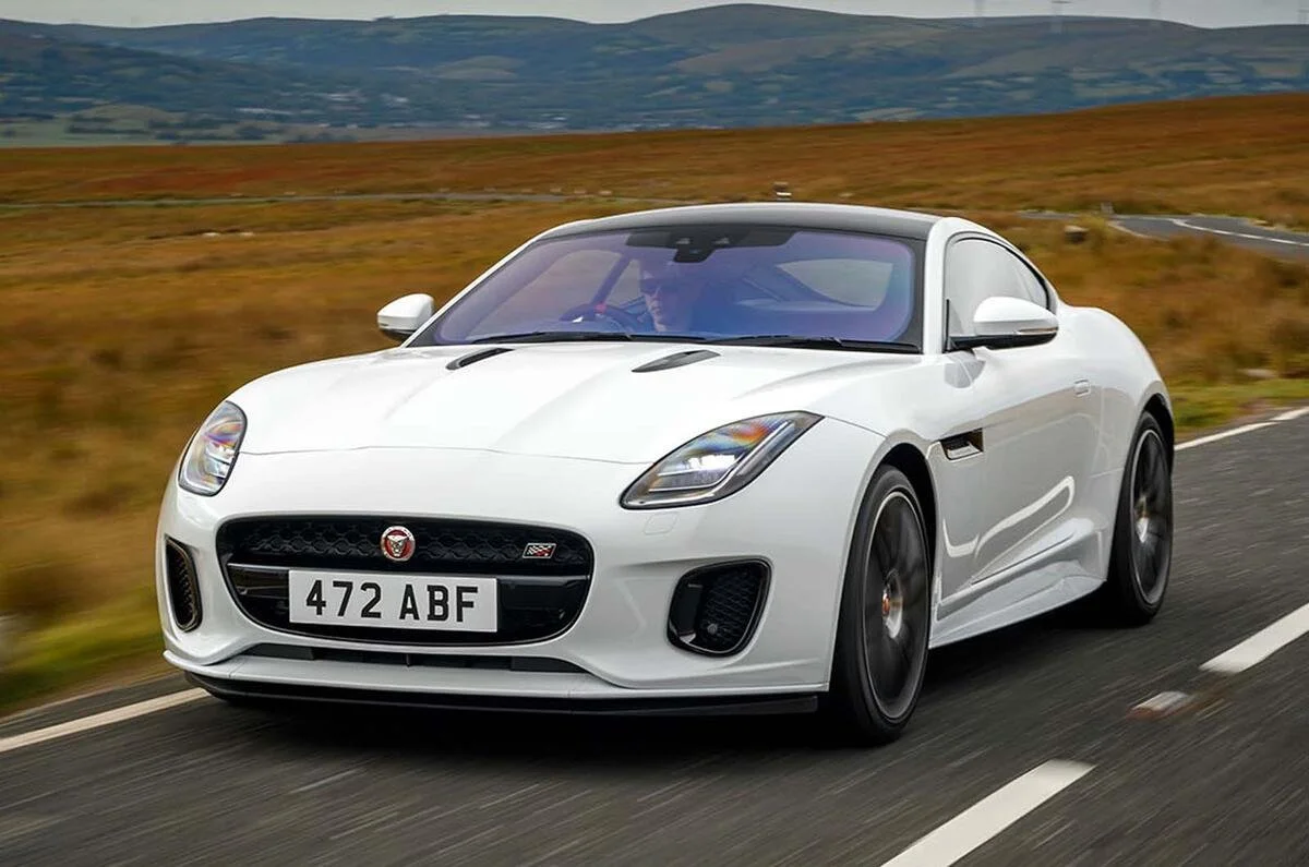 A white Jaguar sports car driving on a rural road with hills and grass in the background.