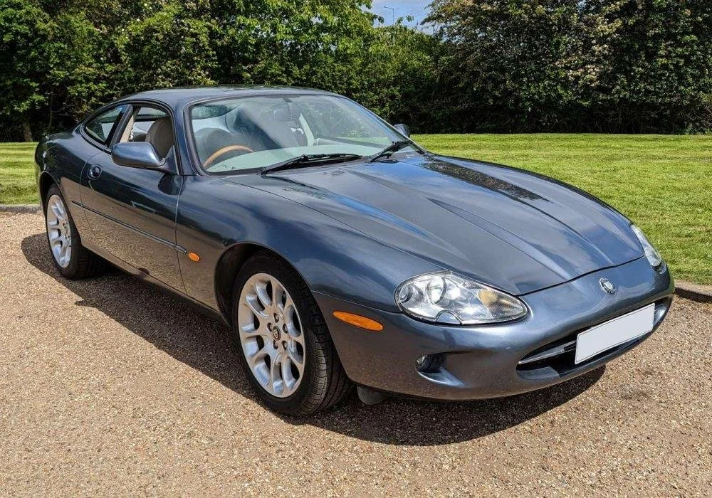 A dark blue Mazda sports car parked outdoors on a gravel surface with greenery and trees in the background.