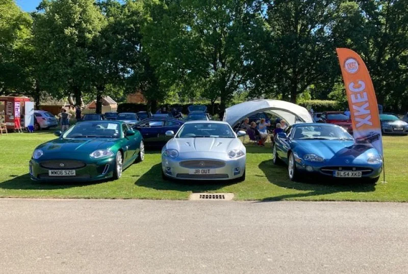 Three vintage sports cars parked on grass at a car show, with a banner and trees in the background.